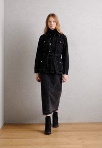Črna jakna iz denima s prilegajočo silhueto, pasom, ki se zapenja, in gumbnimi detajli, v kombinaciji z črno denim krilom in ujemajočimi gležnjači.