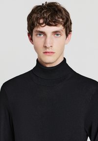 Maglione a collo alto nero realizzato in un tessuto liscio e leggero, con un colletto a coste. Design aderente con maniche lunghe e senza fantasie.