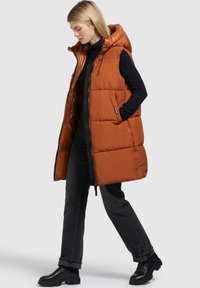 Donna che cammina in dolcevita nera, jeans scuri, stivali neri e un gilet arancione senza maniche con cappuccio, mani in tasca.