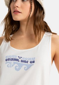 Białe top tank z okrągłym dekoltem, ozdobione niebieskim wzorem graficznym z napisem "Roxy Original Surf Co" oraz wzorami fal.