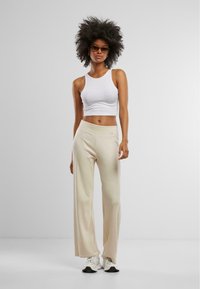 Top corto bianco abbinato a pantaloni a gamba larga di colore crema con vita alta. Modella con occhiali e scarpe da ginnastica, in piedi davanti a uno sfondo neutro.