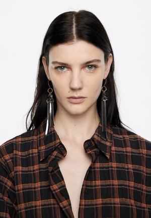 Dlhé čierne náušnice s strapcami a priesvitnými detailmi. Obsahujú zlatý kruhový úchyt. Modelka má na sebe čierno-oranžovú károvanú košeľu.