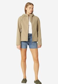 Beige softshell jas met een capuchon, voorrits en zijzakken, gedragen over een wit shirt en denim shorts. Casual schoeisel zichtbaar.