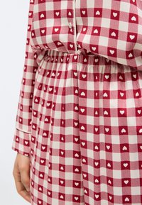 Κόκκινο και άσπρο φόρεμα με καρό μοτίβο και μικρά λευκά καρδούλες, με σούρες στη μέση, κουμπιά και μακρύ μανίκι, εμφανίζεται από τη μέση μέχρι το χέρι.