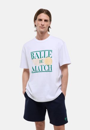 Jeune homme aux cheveux mi-longs portant un t-shirt blanc avec l'inscription « Balle de Match » et un motif de balle de tennis, ainsi qu’un short foncé avec une icône de raquette de tennis.