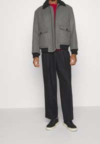 Man som bär en grå zip-up jacka med svarta muddar, svarta vida byxor, svarta sneakers med vita sulor och en röd skjorta under.