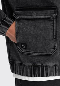 Dettaglio di una giacca di jeans nera che mostra una tasca laterale con patta superiore, bordi cuciti e un'etichetta "STAY REAL" sopra l'orlo. Texture morbida, vestibilità rilassata.