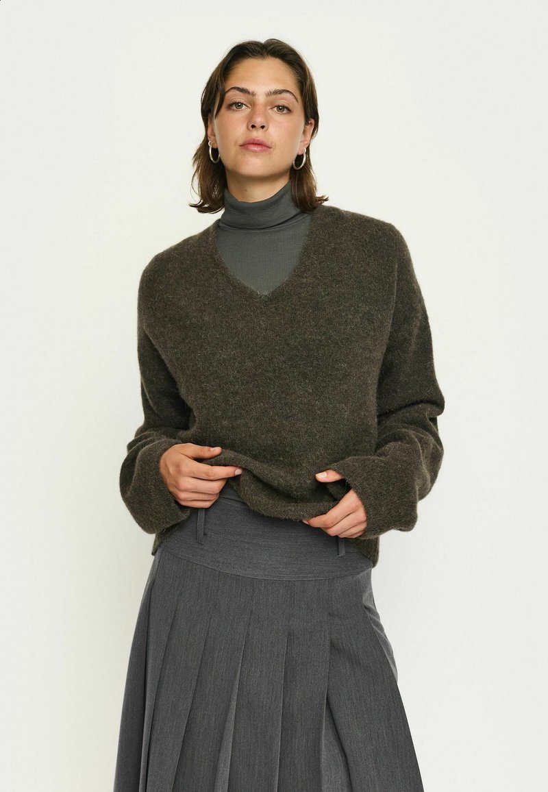 Donna con capelli corti castani che indossa orecchini a cerchio, un maglione scuro felpato a V sopra un dolcevita grigio e una gonna grigia plissettata.