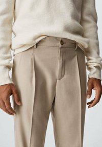 Pantaloni plissettati beige con una vestibilità su misura, realizzati in un tessuto morbido, dotati di chiusura anteriore con bottone e dettagli di cucitura sottili.