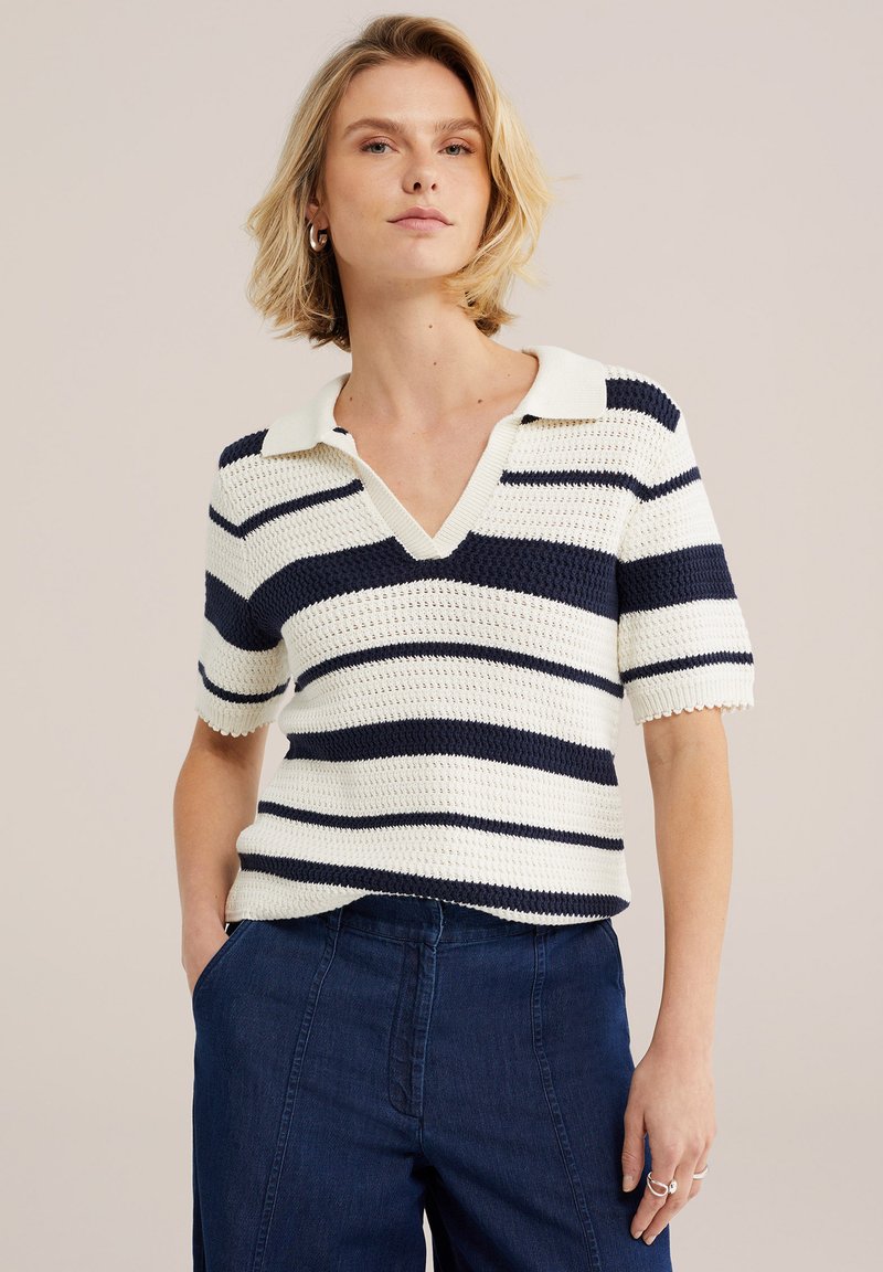 Femme blonde portant un top en maille blanc avec des rayures horizontales bleu marine et un pantalon taille haute bleu foncé, debout avec une main dans la poche.