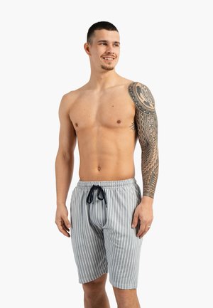 Man zonder shirt met volledige mouw tribal tatoeage, draagt grijs-wit gestreepte short met trekkoord, lacht en kijkt opzij.