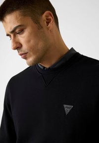 Fekete pulóver kerek nyakkivágással és háromszög logóval a mellrészen, sima anyagból, texturált felülettel.