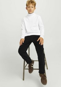 Vit långärmad polotröja, svarta kordbyxor och bruna sneakers. Barn som sitter på en metallpall framför en neutral bakgrund.