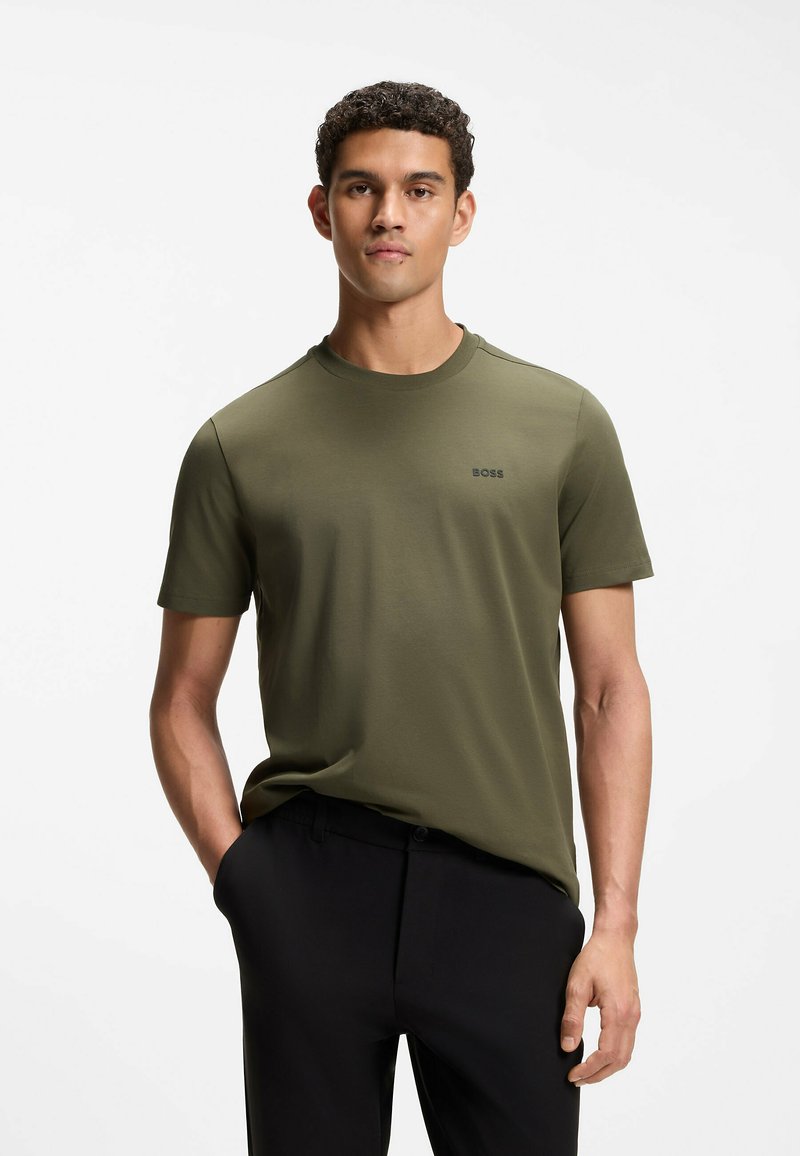 Jeune homme debout avec une main dans la poche, portant un t-shirt BOSS vert olive et un pantalon noir sur fond blanc.