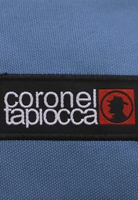 Tela azul con una superficie texturada que presenta una etiqueta negra. La etiqueta tiene texto en blanco que dice "coronel tapiocca" y un logo rojo con un perfil.