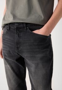 Vaqueros de mezclilla negros de corte ajustado, con un bolsillo frontal, cierre de botón y un leve desgastado para añadir textura. Combinados con una camiseta gris.