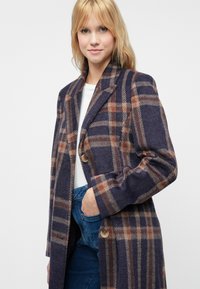 Manteau en laine à carreaux bleu marine, marron et beige. Présente un revers cranté, une fermeture à bouton simple et des poches latérales. Texture lisse, manches longues.