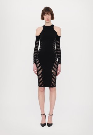 Black, fitted dress with cut-out detailing along the sides and long sleeves, made from a stretchy fabric, paired with black heels.