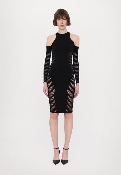 Robe noire ajustée avec des découpes sur les côtés et des manches longues, fabriquée dans un tissu extensible, assortie à des talons noirs.