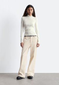 Langärmliges, hellcremefarbenes Oberteil mit Rüschenausschnitt, kombiniert mit weit geschnittenen, gerippten beigen Hosen und schwarzen Plateauschuhen.