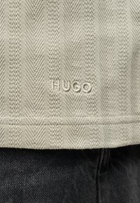 Κοντινό πλάνο από μπεζ υφασμάτινη επιφάνεια με κάθετα σχέδια Chevron και πλεγμένα μοτίβα, κεντημένα με τη λέξη "HUGO" κοντά στο κάτω μέρος πάνω από σκούρο τζιν.