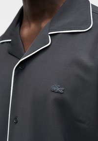 Chemise grise à manches courtes avec un liseré blanc, arborant un logo crocodile noir brodé sur la poitrine gauche, fabriquée en tissu lisse.