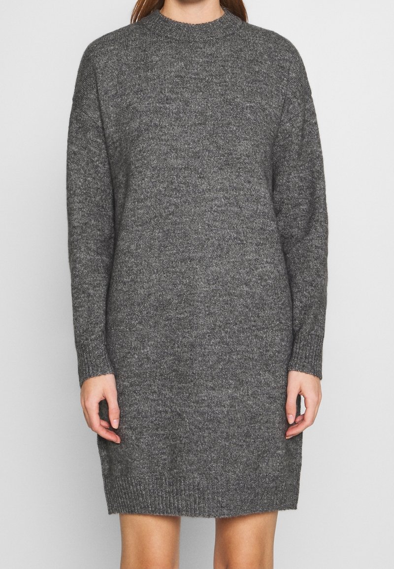 Femme portant une robe pull longue en mailles gris foncé, manches longues, longueur au genou, avec un col rond, sur un fond clair uni.