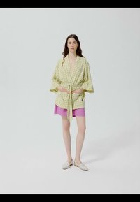 Femme portant une veste ceinturée à carreaux vert clair sur une jupe rose, debout avec les mains dans les poches devant un fond blanc uni.