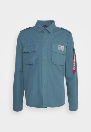 Cămașă din bumbac albastru cu mânecă lungă, cu epauleți pe umeri, două buzunare în față și un etichetă roșie cu text pe manșetă. Design cu închidere cu nasturi pe față.