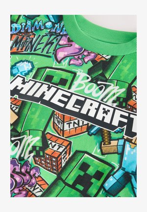 Žalias medvilninis marškinėlis su Minecraft dizainu, vaizduojančiu pikselių meno personažus, žodžius „Boom“ ir „Minecraft“ ryškia juoda spalva spalvingame fone.