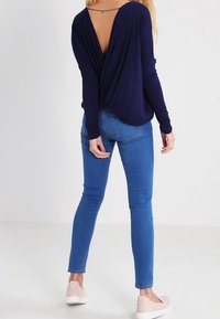 Nainen, jolla on tummansininen pitkähihaista paitaa syvällä V-leikkauksella takana, siniset skinny-farkut ja vaaleanpunaiset liukuvaatteet, seisoo valkoisella lattialla.