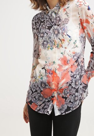 Chemisier à manches longues boutonné avec un motif floral en noir, blanc, orange, avec des touches de bleu et de vert, porté avec un pantalon noir.