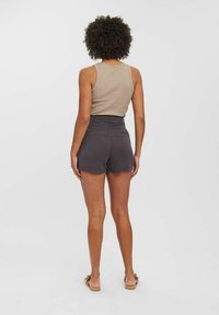 Femme aux cheveux bouclés portant un débardeur beige, un short taille haute gris foncé et des sandales plates beige, debout de dos devant un fond uni.