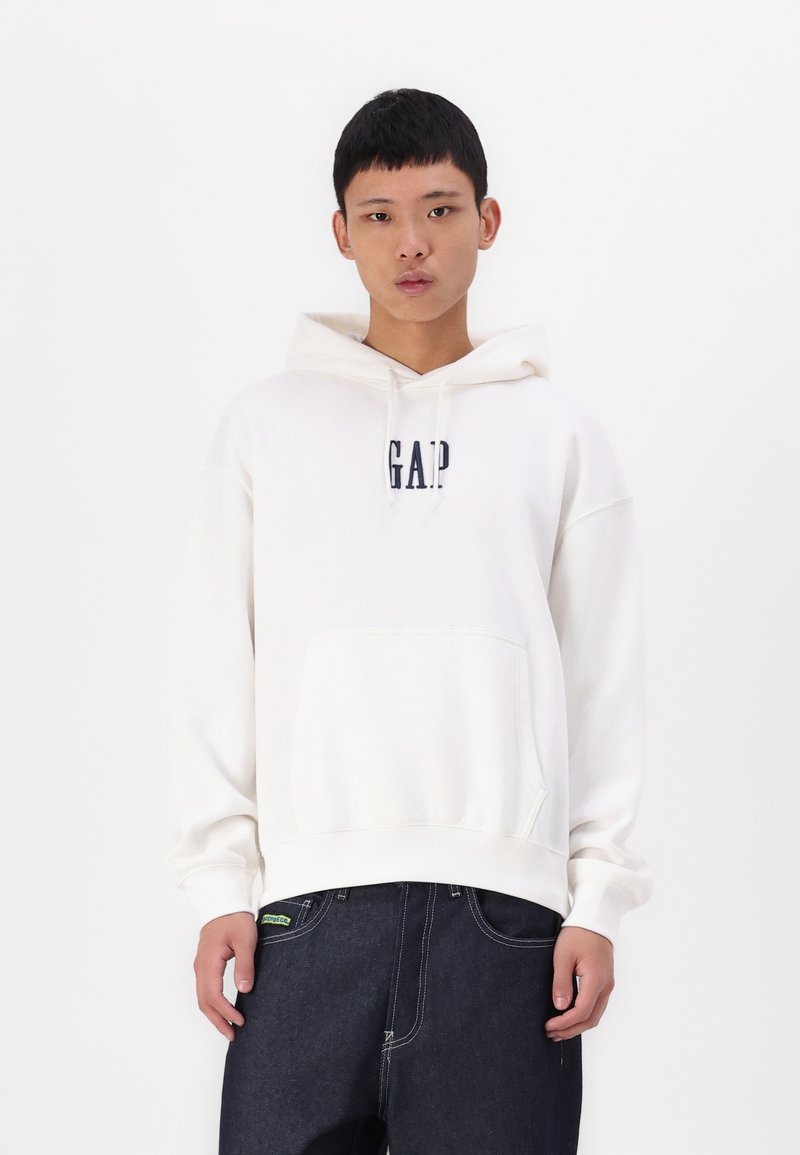 Sweatshirt à capuche blanc avec le logo "GAP" en marine, poche kangourou, manches côtelées et coupe décontractée, associé à un jean en denim foncé.