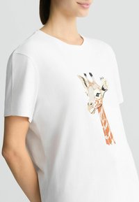 T-shirt en coton blanc avec un motif de girafe en orange, marron et noir, à manches courtes et avec un col rond.