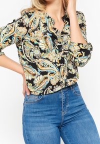 Svart skjorta med färggomt paisley-mönster, knäppning framtill och uppvikta ärmar, tillsammans med blå denimjeans. Blandad bomullsframställning.