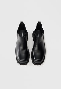 Mustat nahkaiset nilkkurit, joissa on sileä pinta, joustavat sivuinsertit ja paksut kumipohjat. Minimalistinen muotoilu ja vetokahvat päällä.