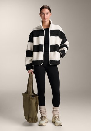 Femeie purtând o jachetă fleece în dungi alb-negru, colanți negri, încălzitoare bej pentru picioare și adidași, ținând o geantă mare de culoare verde măsline.