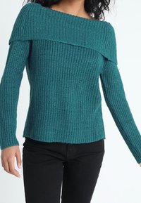 Pull turquoise en maille à épaules dénudées avec texture côtelée, manches longues et ourlet droit, associé à un pantalon noir.