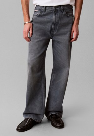 Mann trägt locker sitzende graue Jeans, schwarze Loafers, ein weißes Hemd und ein silbernes Armband, steht vor einem einfachen hellen Hintergrund.