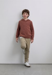 Maglione con cappuccio color ruggine con polsini a coste, abbinato a pantaloni beige e sneakers bianche. Parete liscia sullo sfondo.