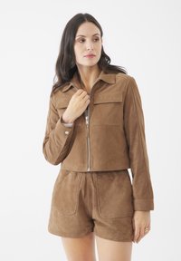 Giacca cropped in suede marrone con zip argentata, due tasche frontali e maniche lunghe. Abbinata a pantaloni corti in suede marrone coordinati.