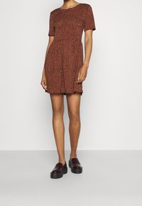 Lyhythihainen mekko oranssinruskeana, mustalla leopardikuvioinnilla, keskeltä vyötäröltä kapeana ja levenevällä helmalla. Yhdistetty mustiin kiiltäviin korokepohjakenkiin.