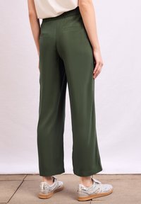 Groene, brede broek met een gladde textuur. Heeft een elastische tailleband en twee achterzakken. Draagt een paar zilveren sneakers.