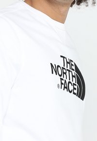 Man som bär en vit sweatshirt med en svart "The North Face"-logotyp på bröstet, visad från nacken till mitten av överkroppen.