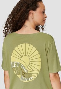 Olivenfarbene Kurzarm-T-Shirt mit einem großen gelben Sonnenmotiv und dem Text "LASS DAS SONNENSCHEIN IN DEIN HERZ" in Gold. Glatte Textur.