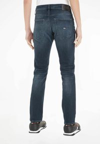 Jeans de mezclilla azul oscuro con corte Slim, cinco bolsillos y un parche de cuero en la cintura. Llevados con zapatillas deportivas negras.