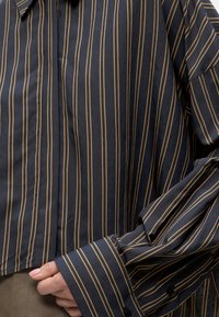 Chemise bleu marine à manches longues avec fines rayures verticales beige, poignets boutonnés, et une main partiellement rentrée dans le devant de la chemise.
