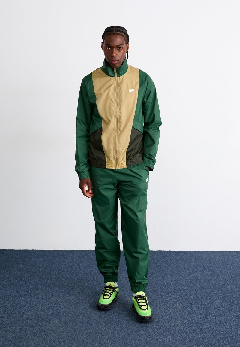 Zeleno-bež trenirka iz gladkega materiala, ki ima elastični pas, jakno na zadrgo in svetlo zelene športne copate s črnimi detajli.