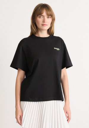 T-shirt nera oversize in cotone con maniche corte. Presenta un piccolo logo verde sul lato sinistro del petto. Design semplice senza ulteriori motivi o dettagli.